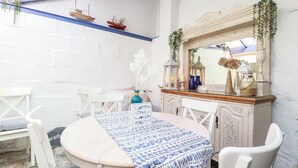 Een eetgedeelte met een ronde tafel, witte stoelen, een blauwe tafelkleed met patroon en een decoratieve spiegel erboven.