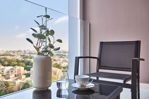 A balcony with a view of a cityscape, a table with a glass of water, a cup, and a vase with green plants. - Modern 1BR | Golf Views | Vista Dubai Sports City (Dubai)