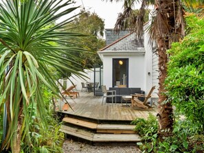 A wooden deck with outdoor furniture, a white building with a dark door, and surrounding greenery. - Riverside Cottage With Estuary Views (Fowey)