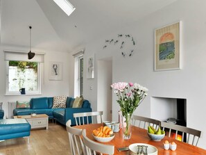 A modern living room with a blue sofa, a dining table with a vase of flowers, and a fireplace. - Riverside Cottage With Estuary Views (Fowey)
