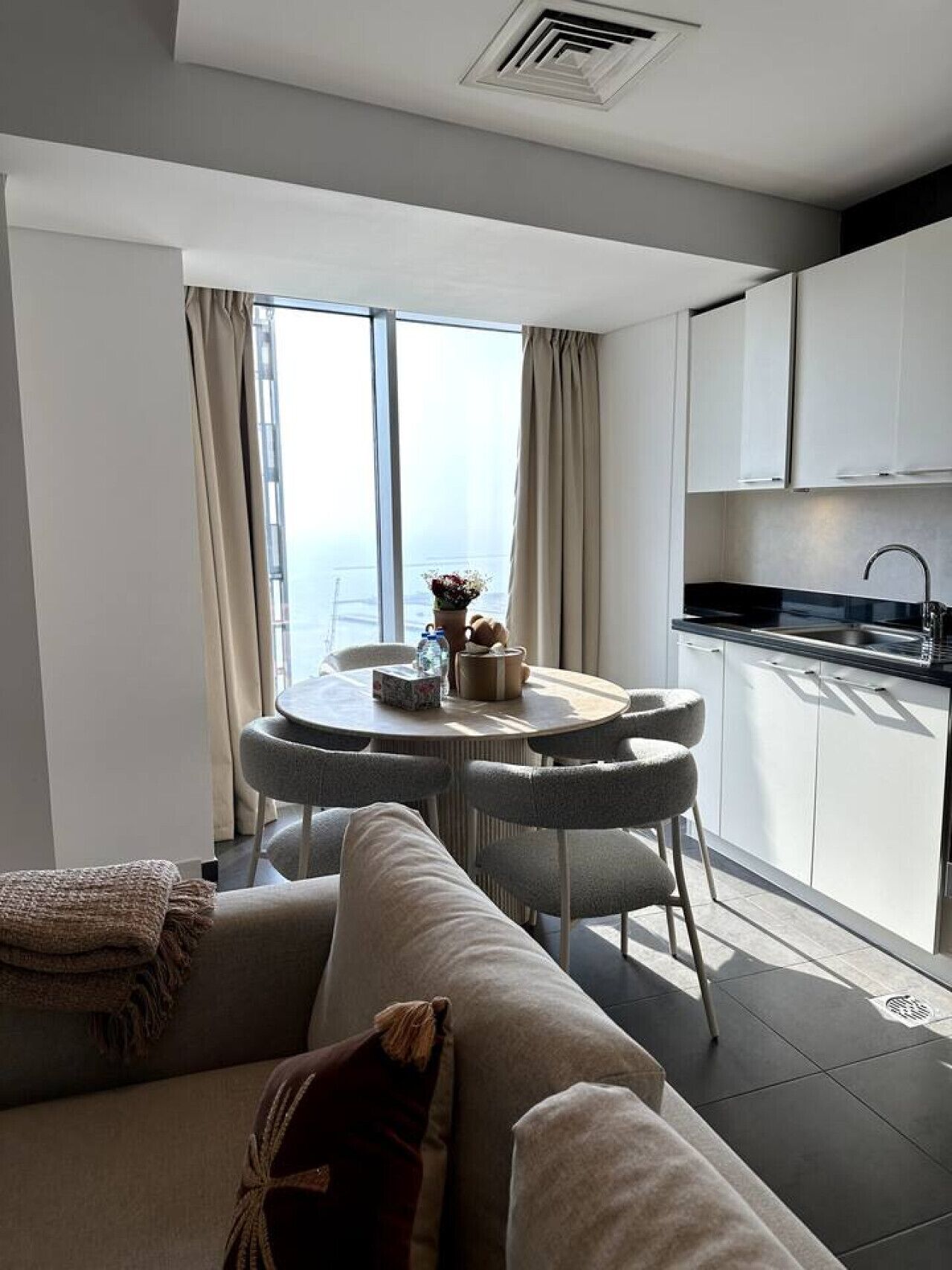 Een moderne woonkamer met een bank, een ronde eettafel met stoelen, een keuken met kasten, en een groot raam met gordijnen.