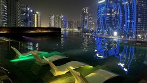 Un paisaje urbano nocturno con edificios iluminados y un estanque reflectante con mobiliario moderno.