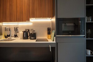 A modern kitchen with wooden cabinets, a built-in microwave, a kettle, and various utensils. - Accessible Studio | Long Stay Ideal | StayCozy (London)
