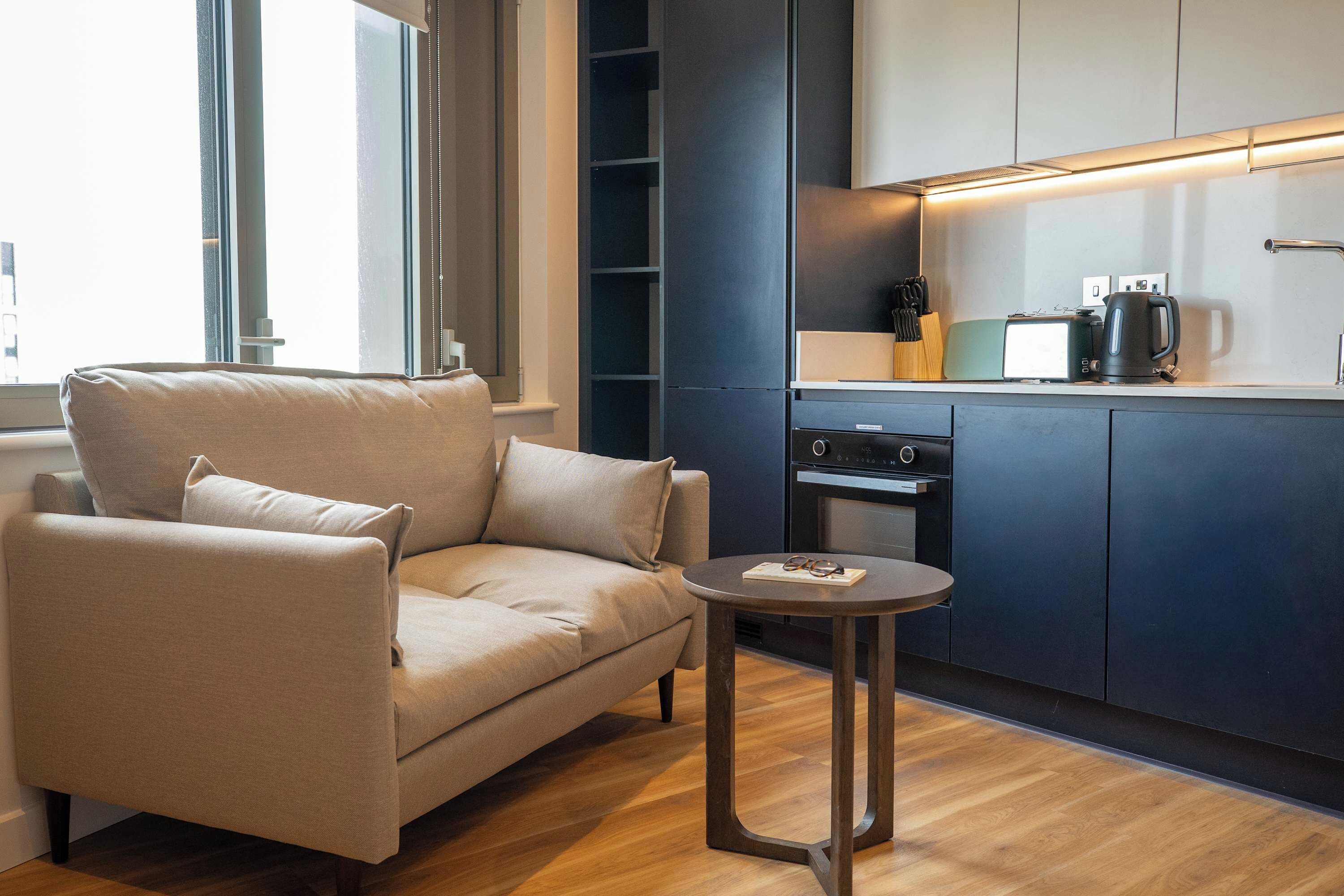 A modern living room with a sofa, a small round table, and a kitchen area with cabinets and appliances.