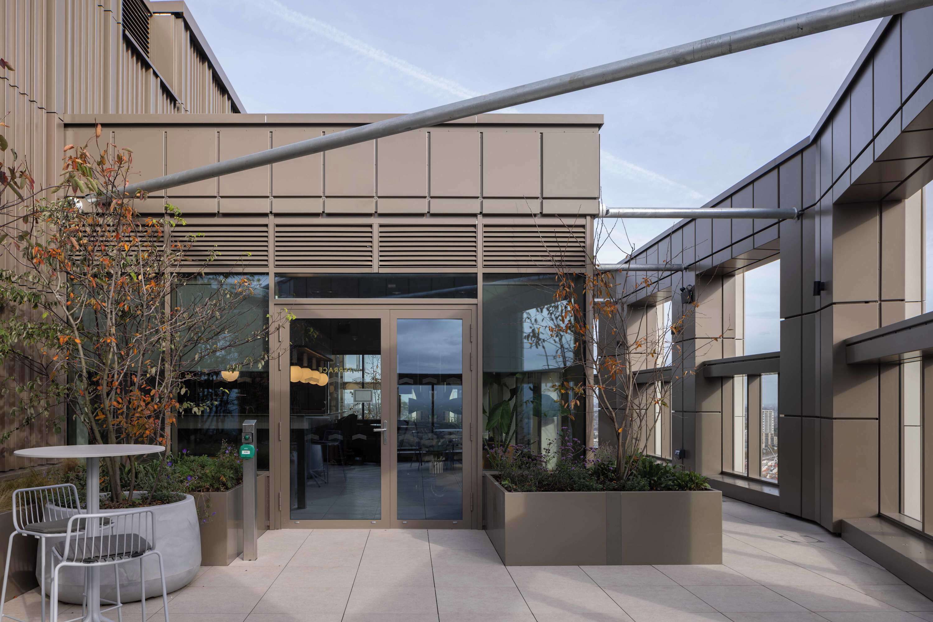 A modern building entrance with a glass door, outdoor seating, and planters.