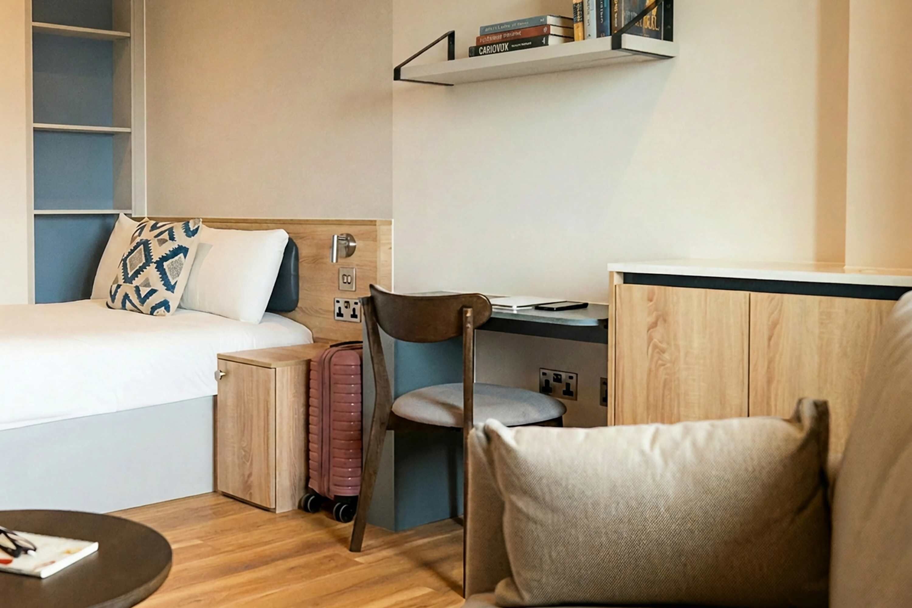 A compact hotel room with a bed, a desk, a chair, a small cabinet, and a suitcase.