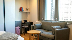 Ein modernes Hotelzimmer mit grauem Sofa, einem kleinen Holztisch, einem Schreibtisch mit Laptop, einem pinkfarbenen Koffer und einem Bücherregal mit Büchern und einem Bilderrahmen.