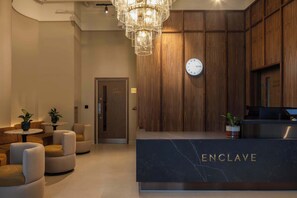 A reception area with a marble counter labeled 'ENCLAVE,' wooden paneling, a clock, and a chandelier. - Contemporary Studio | Cowork + Gym | StayCozy (London)