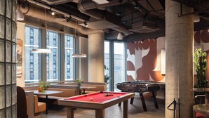 Una mesa de billar, zona de estar y una mesa de fútbol de mesa en un espacio interior moderno con ventanas amplias y elementos de diseño industrial.