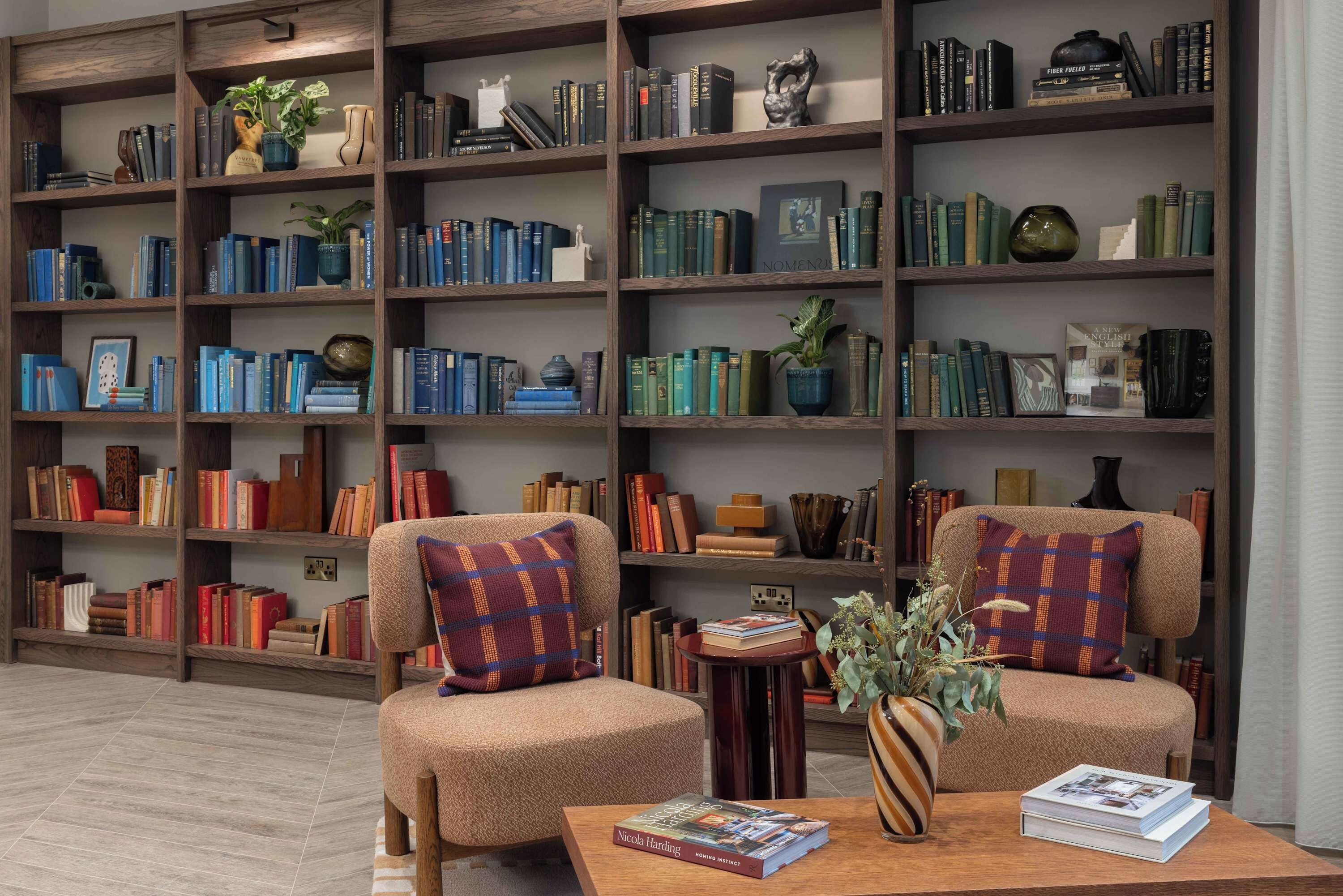 A library with multiple bookshelves, two armchairs, and a coffee table with books and a vase.