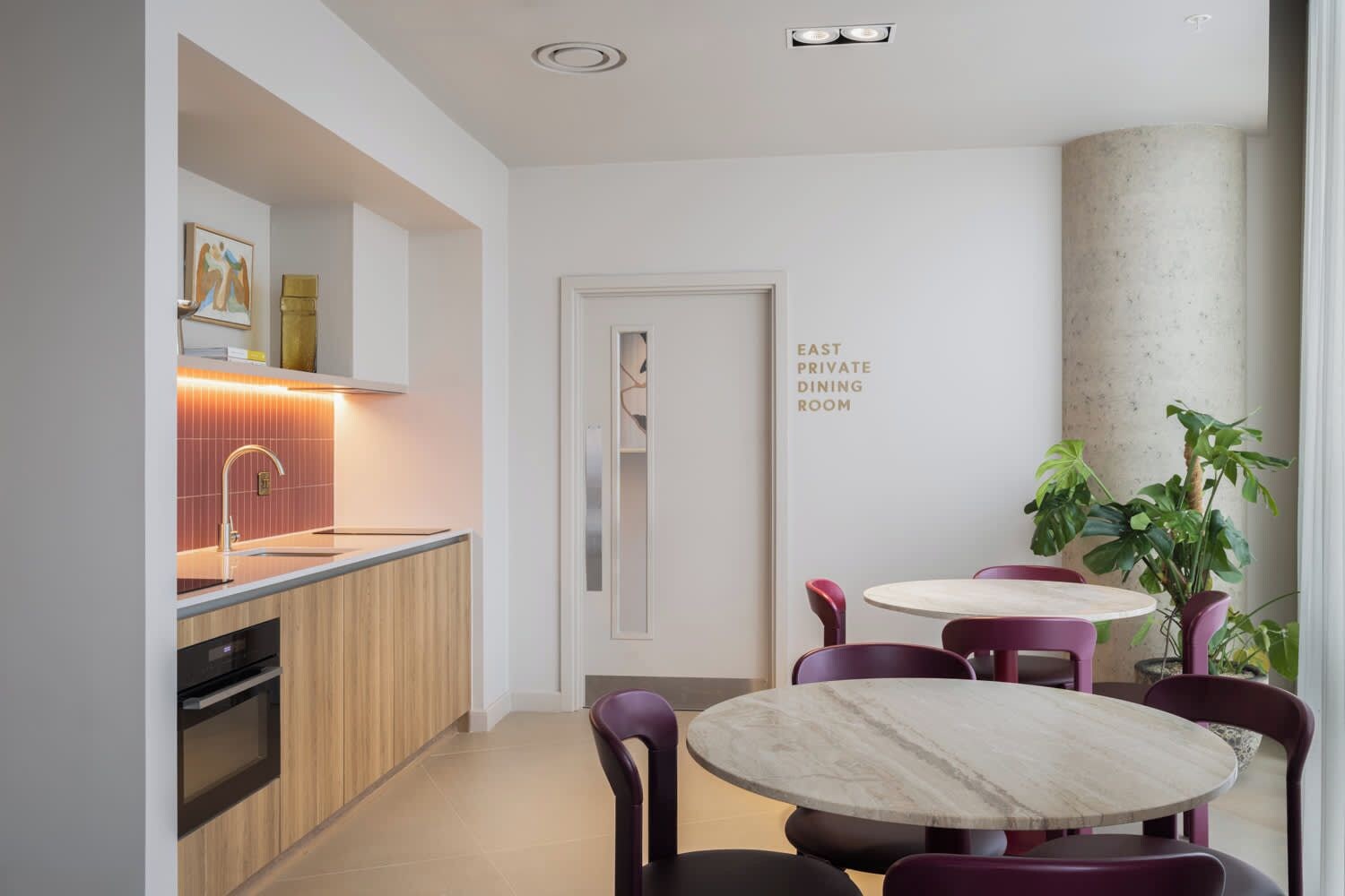 A modern kitchen with a built-in oven, a sink, and a countertop with a lighted backsplash. There is a dining area with a round table and purple chairs.
