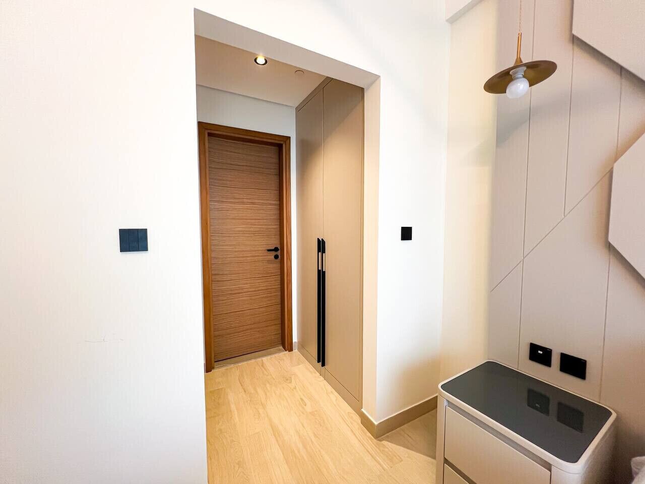 Ein Korridor mit einer Holztür, einem schwarzen, vertikalen Gegenstand und einem kleinen, schwarzen Quadrat auf der Wand.