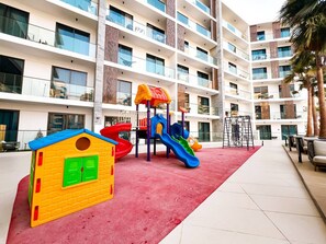 Ein Spielplatz mit Rutschen und einer Kletterkonstruktion, umgeben von einem Gebäude mit Balkonen und einem roten Teppich.
