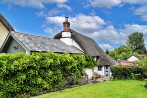 A traditional thatched-roof house with a white exterior, surrounded by lush greenery and a clear blue sky. - Cottage With Inglenook Hearth (Winkleigh)
