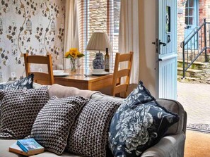 Ein gemütliches Zimmer mit einem Holztisch, zwei Stühlen, einer Couch mit gemusterten Kissen, einer Vase mit Sonnenblumen und einer Lampe.