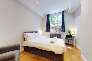 A hotel room with a bed, a desk, a chair, a window with curtains, and a radiator. - Modern 2BR w/patio, Walk to Westminster/Buckingham (London)