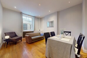 A modern living room with a sofa, a dining table, and chairs. There is a window with curtains, a radiator, and a clock on the wall. - Modern 2BR w/patio, Walk to Westminster/Buckingham (London)