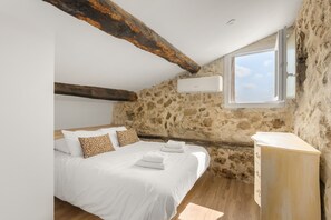 Ein Schlafzimmer mit schrägen Wänden, freiliegenden Holzbalken, einer Steinwand, einem Bett mit weißer Bettwäsche und gemusterten Kissen, einem Nachttisch und einem Fenster mit Blick auf den Himmel.