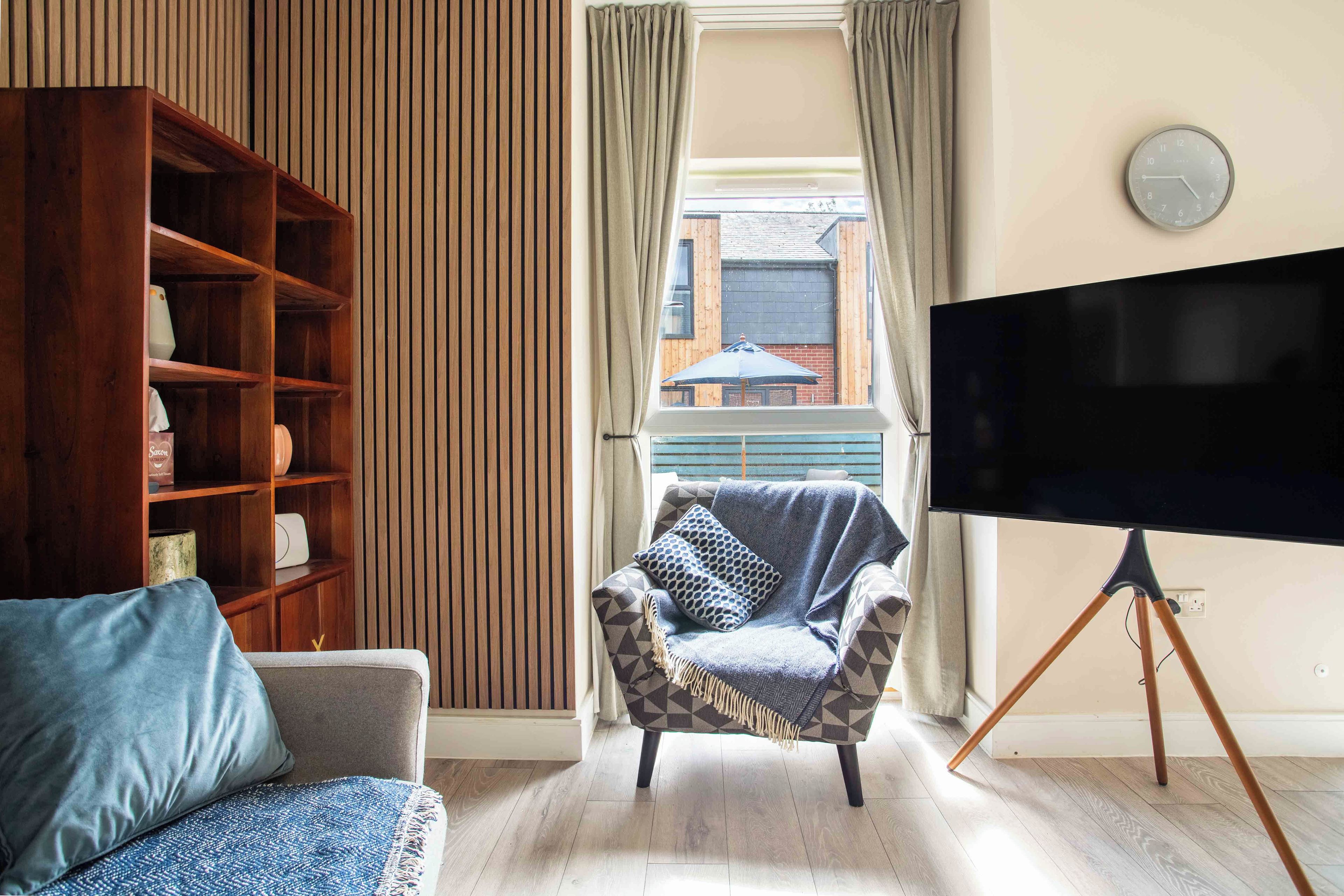 A living room with a sofa, armchair, TV on a stand, wooden bookshelf, and a window with curtains.