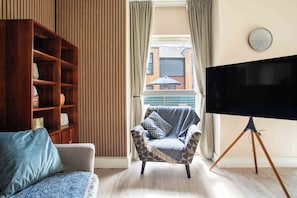 Ein Wohnzimmer mit einer Couch, einem Sessel, einem Fernseher auf einem Schrank, einem Holzregal und einem Fenster mit Vorhängen.