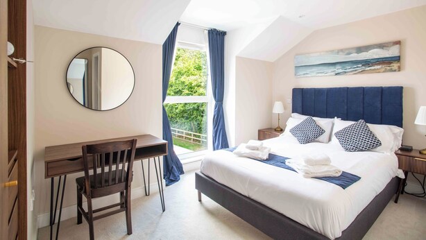 Un dormitorio con cama, escritorio, silla, espejo, ventana con cortinas, mesita de noche, lámpara y un cuadro en la pared.