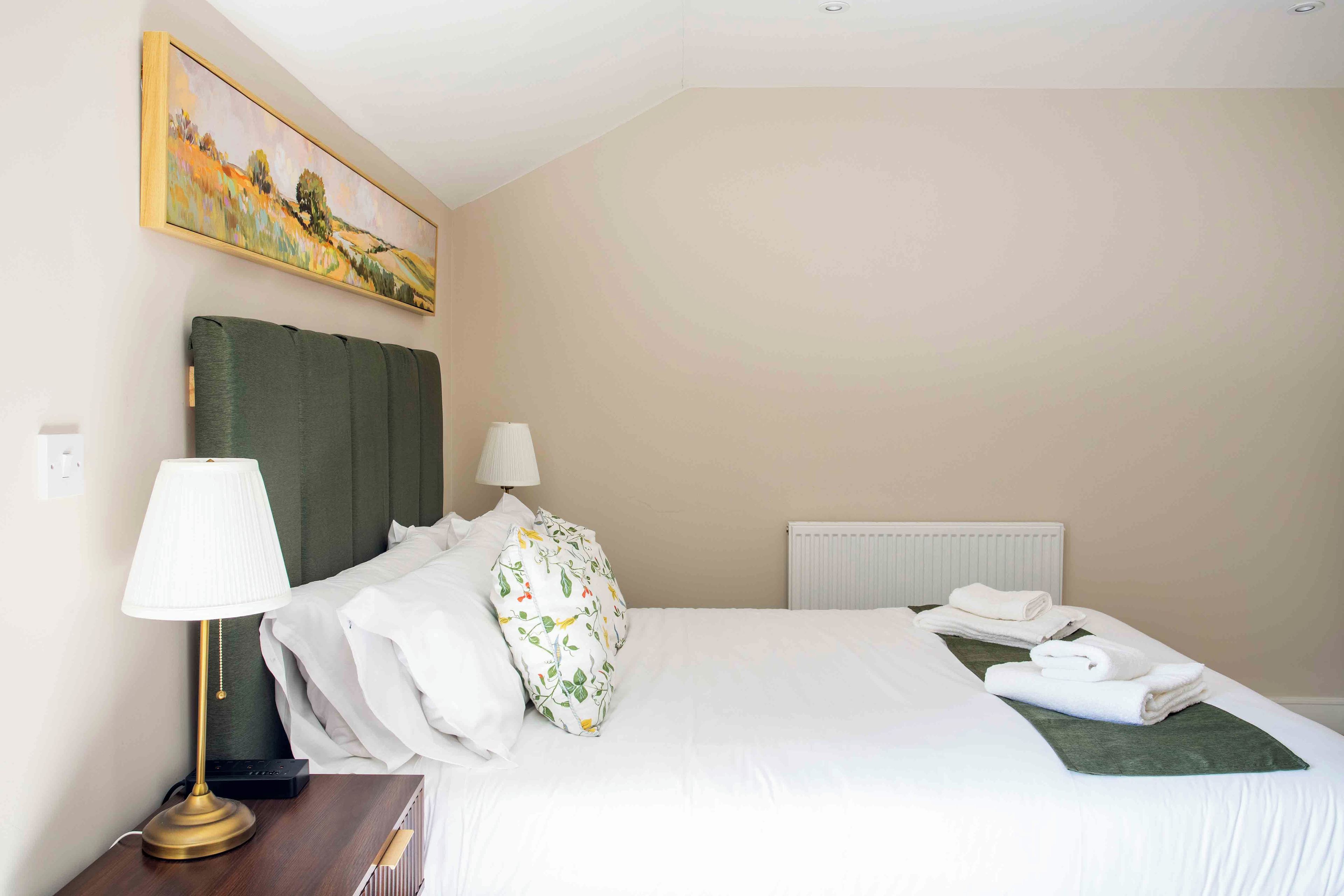 A hotel room with a bed, two bedside lamps, a painting on the wall, and a radiator.