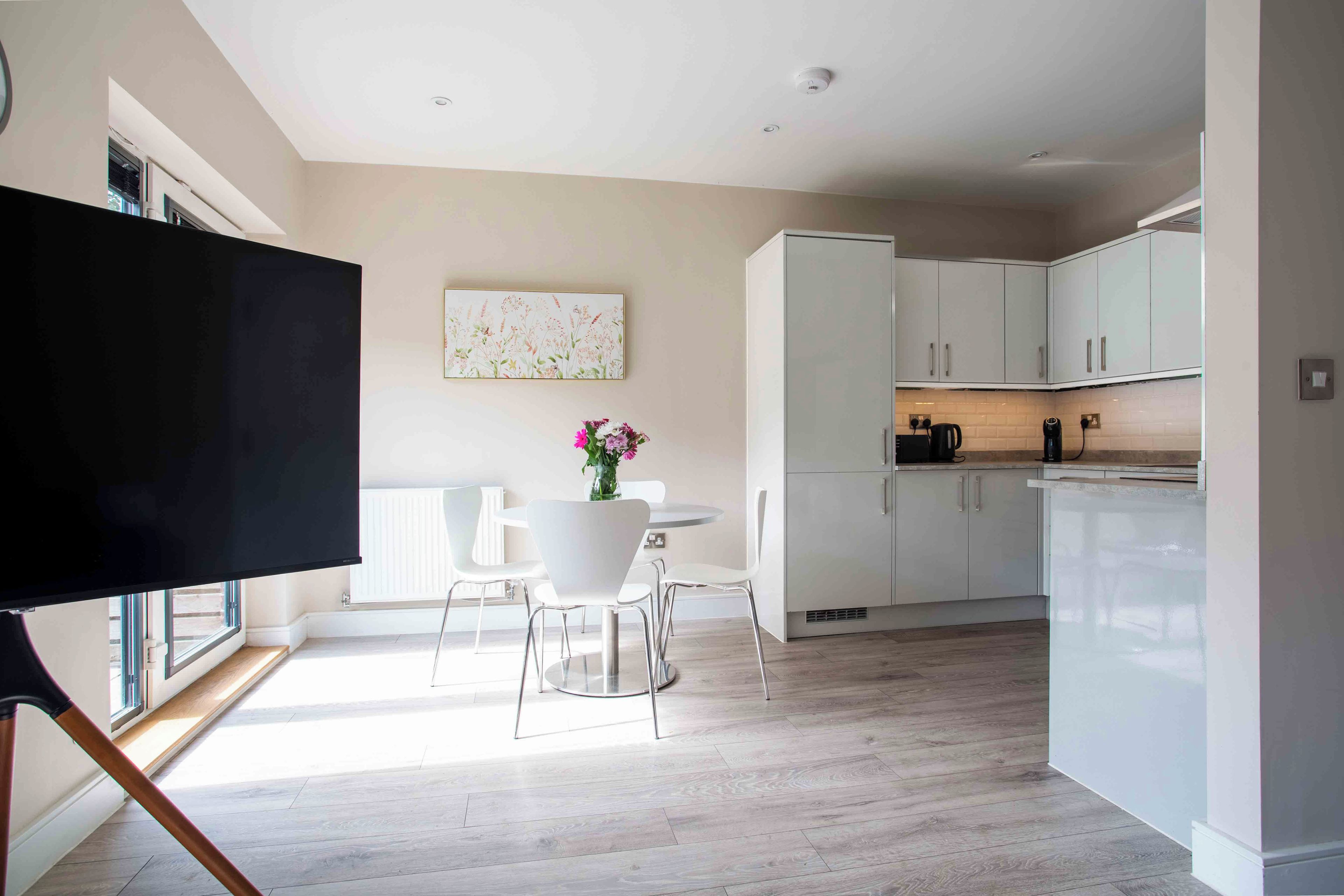 A modern kitchen with white cabinets, a dining area with a white table and chairs, and a flat-screen TV mounted on a wall.
