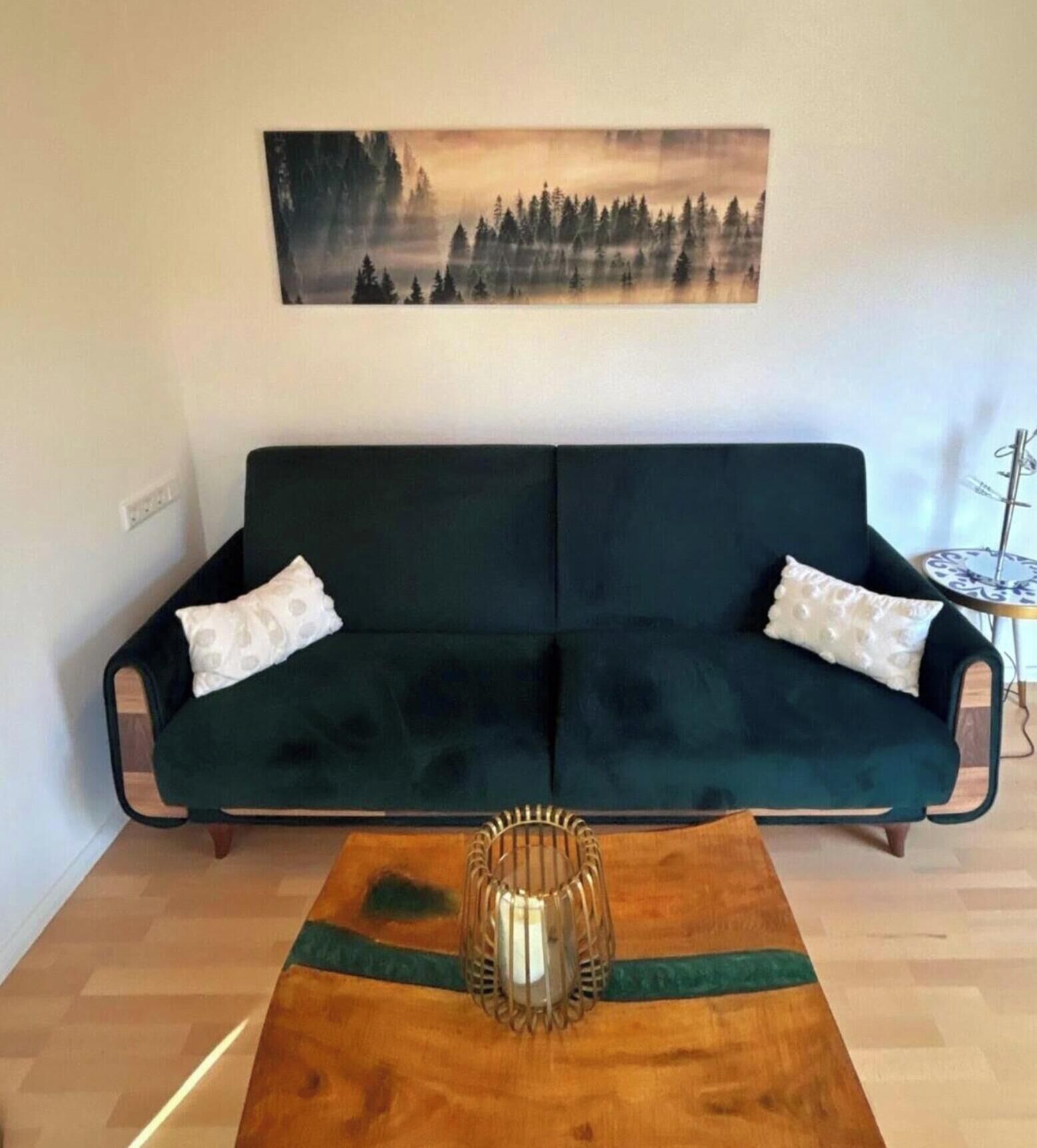A room with a dark green sofa, two white pillows, a wooden coffee table with a candle, and a painting of a forest on the wall.