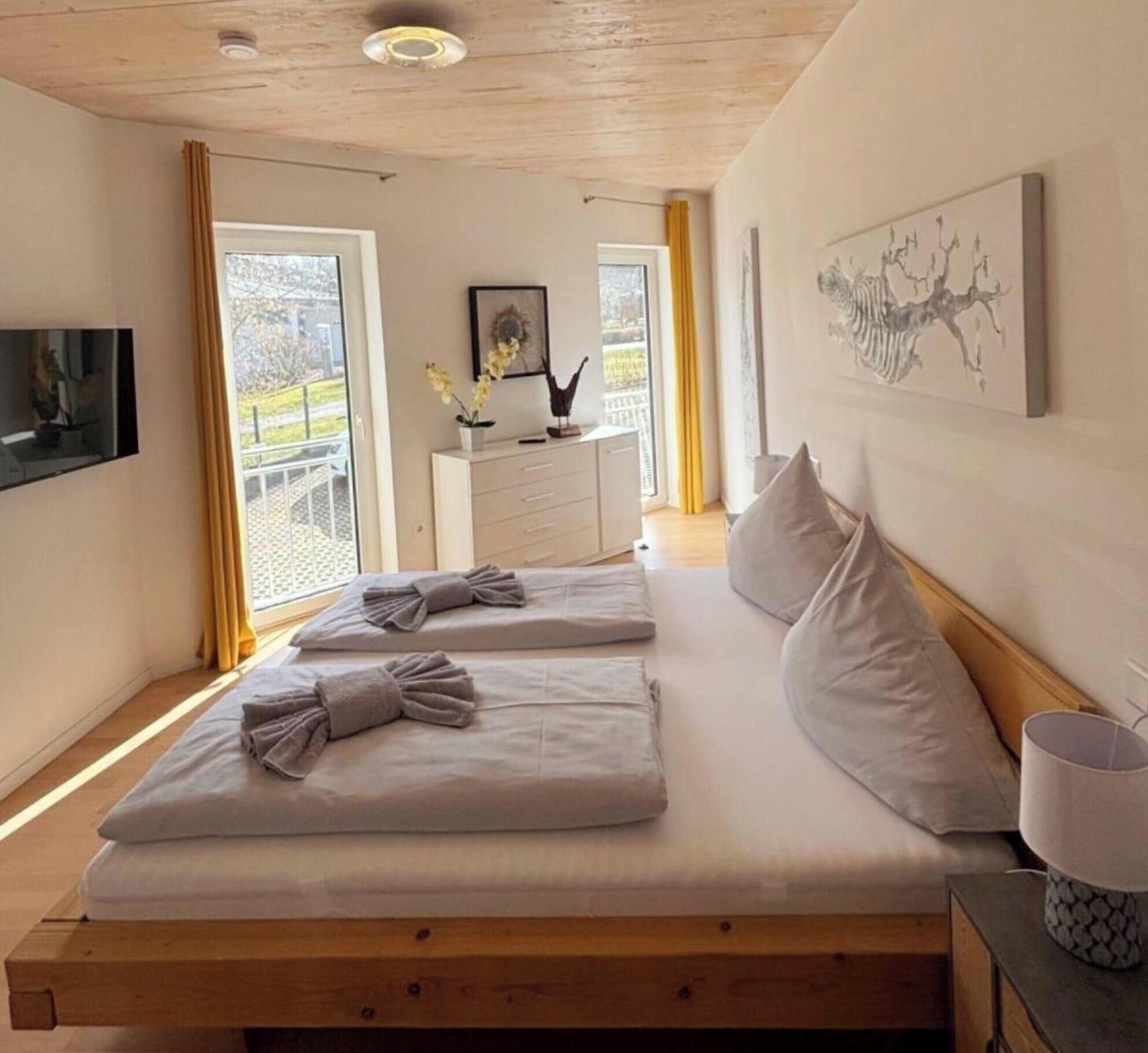 A bedroom with a wooden bed, two pillows, a bedside table with a lamp, a dresser, and a painting on the wall.