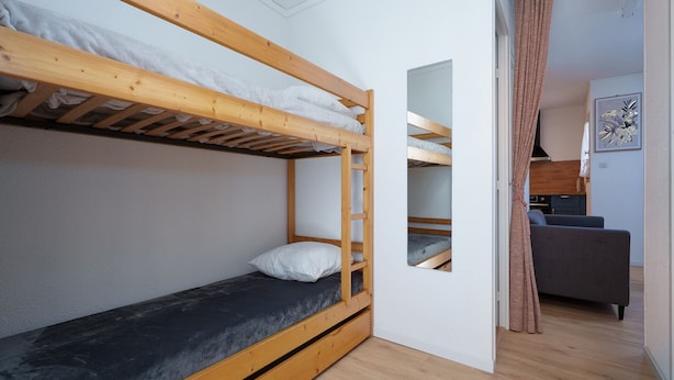 Ein Schlafraum mit Etagenbetten, Holzbalkendecke, einer grauen Couch und einer sichtbaren Küchenzeile durch einen offenen Durchgang.