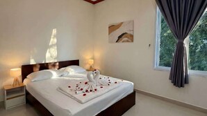 A modern hotel room with a large bed, a nightstand, a bedside lamp, a wall-mounted light fixture, a window with curtains, and a painting on the wall. - Srithanu Villa (Tambon Ko Pha-ngan)