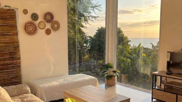 A modern living room with a sofa, coffee table, and a large window offering a view of the outdoors. - Srithanu Villa (Tambon Ko Pha-ngan)