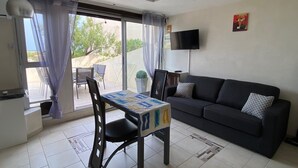 Ett vardagsrum med en svart soffa, ett bord med ett mönstrat bordsduk, en tv som är monterad på väggen och en utsikt genom fönstret.