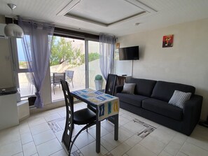 A living room with a black sofa, a table with a patterned tablecloth, a TV mounted on the wall, and a view through the window. - Air-conditioned apartment, sea view - 4 guests (Port Leucate)