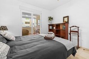 A bedroom with a bed, a wooden bookshelf, a chair, a lamp, and a view through a sliding door. - Apartment with a Terrace and Swimming Pools in El Puerto (Estepona)