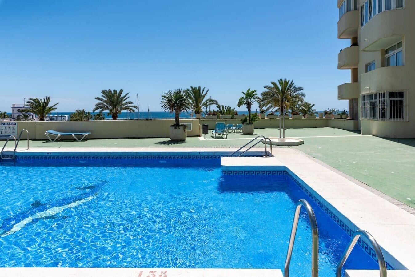 A swimming pool with a clear blue water, surrounded by lounge chairs and palm trees, with a building in the background.