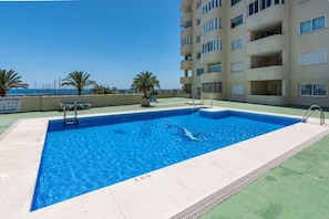 A rectangular swimming pool with a clear blue water surface, surrounded by a low-rise building with balconies, and a clear sky in the background. - Apartment with a Terrace and Swimming Pools in El Puerto (Estepona)