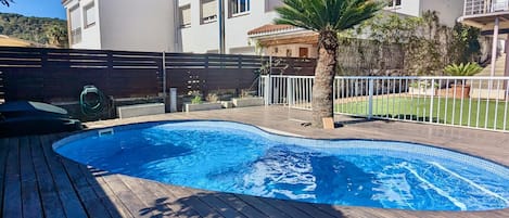 Una piscina con cubierta de madera, rodeada por una valla blanca, y una casa con techo de tejas al fondo.