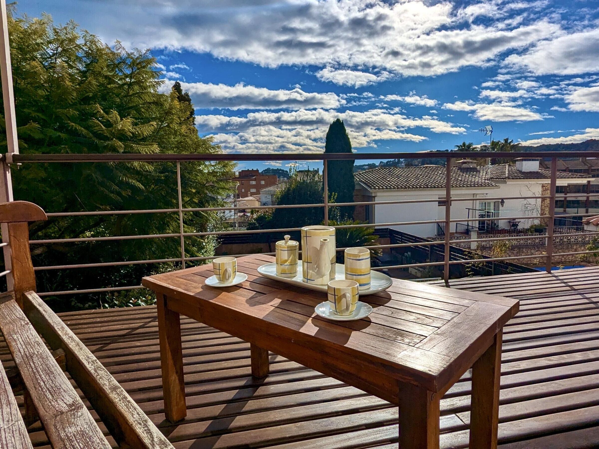 Una terraza de madera con una mesa preparada para dos, con vistas a una zona residencial y un cielo despejado.