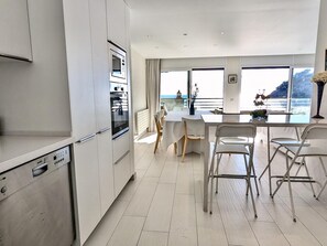 A modern kitchen with a built-in oven and dishwasher, a dining area with a table and chairs, and a view of the sea through large windows. - Spacious apartment, panoramic view - 9 guests (Tossa de Mar)