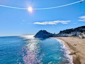 A coastal beach scene with a clear sky, a sun reflecting on the water, and a rocky outcrop. - Spacious apartment, panoramic view - 9 guests (Tossa de Mar)