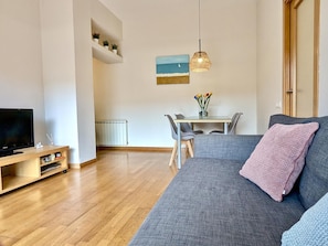 Ein Wohnzimmer mit Sofa, Fernseher, Esstisch mit Stühlen, einer Pendelleuchte und einem Bild an der Wand.