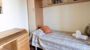 Ein kleines Zimmer mit einem Einzelbett, einem Holztisch, einem Regal mit Dekoration und einer bettenbezogenen Tagesdecke mit Muster.