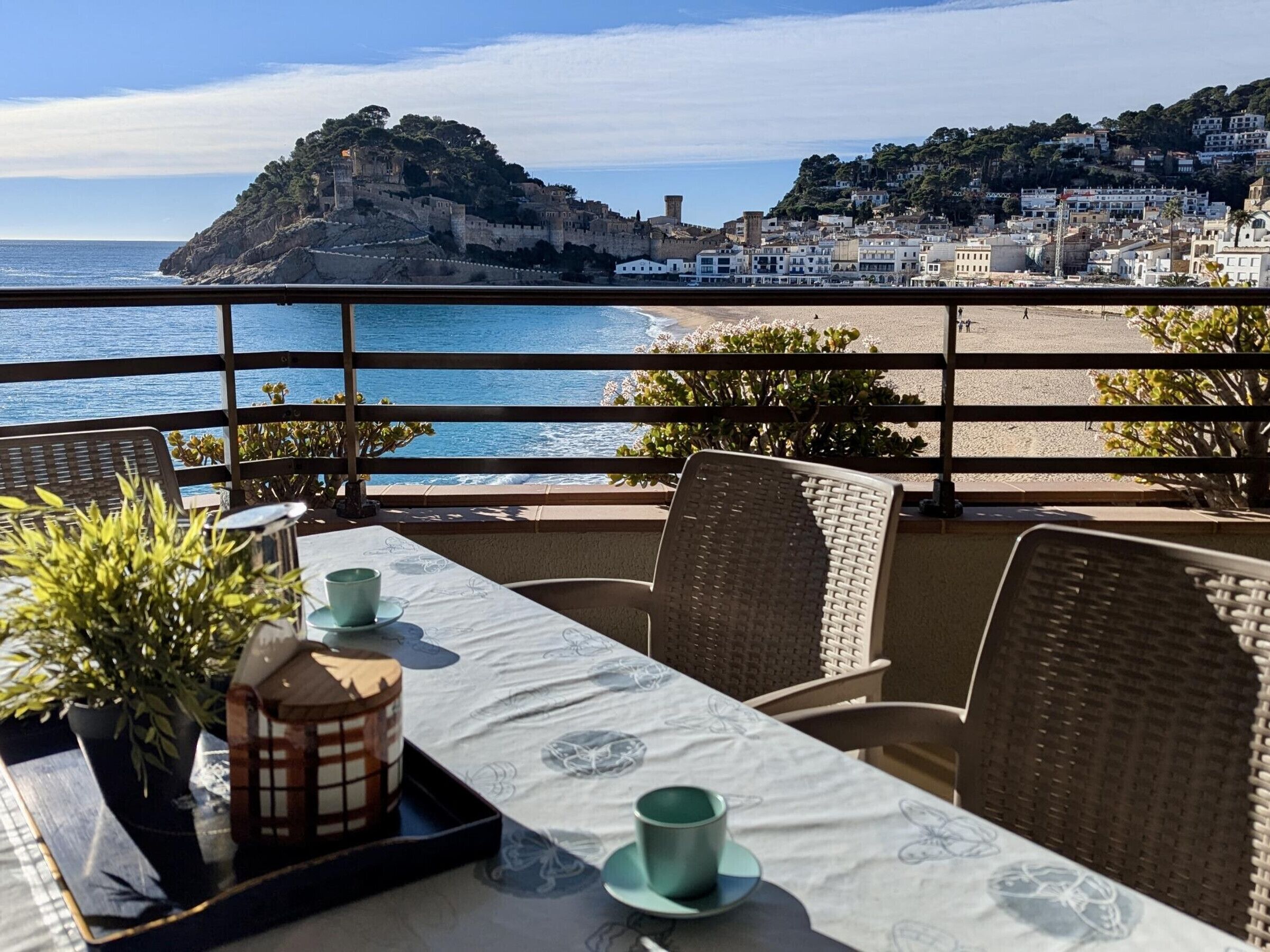 A terrace with a table set for two, overlooking a beach and a historic hilltop structure.