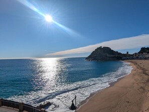 Ein Strand mit blauem Himmel, der Sonne auf dem Wasser und einem Felsen mit Gebäuden.