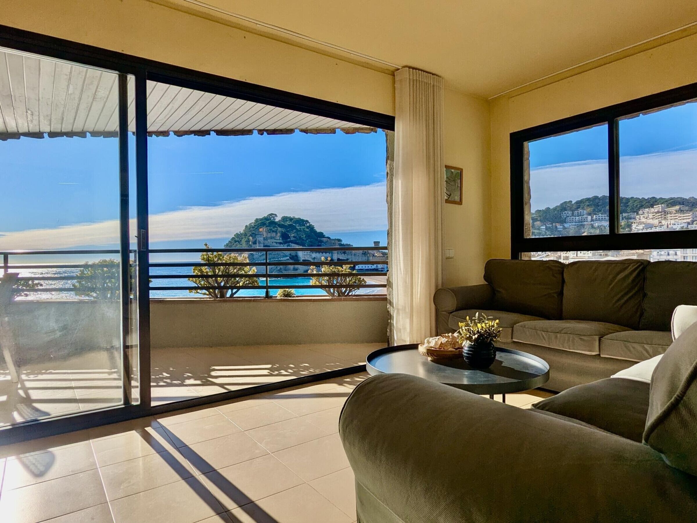 A spacious living room with a large balcony, a sofa, a coffee table, and a view of a coastal landscape.