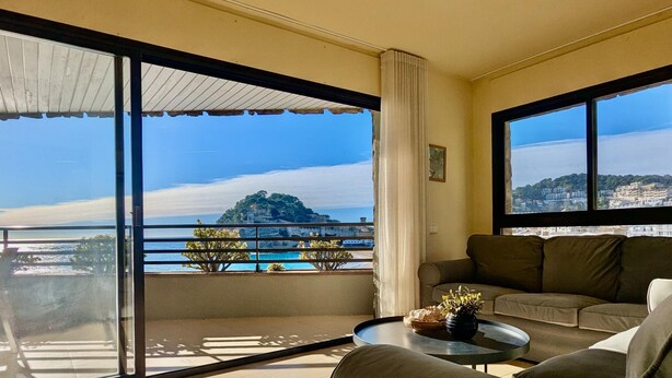 Un soggiorno spazioso con un ampio balcone, un divano, un tavolino da caffè e vista su un paesaggio costiero.