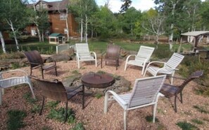 An outdoor seating area with various chairs and a round table, surrounded by trees and a building in the background. - Lazy Duck Cottages The Whole Flock In One Booking (Duck Creek Village)
