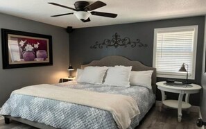 Un dormitorio con cama, ventilador de techo, mesita de noche y ventana con persianas.