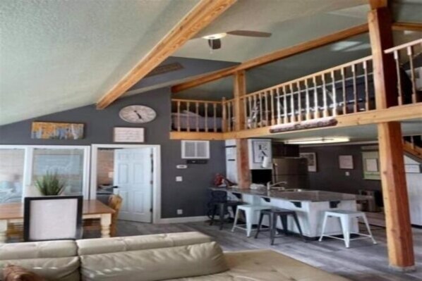 A spacious living area with a high ceiling, a kitchen with bar stools, and a balcony with a railing. - Lazy Duck Cottages The Whole Flock In One Booking (Duck Creek Village)